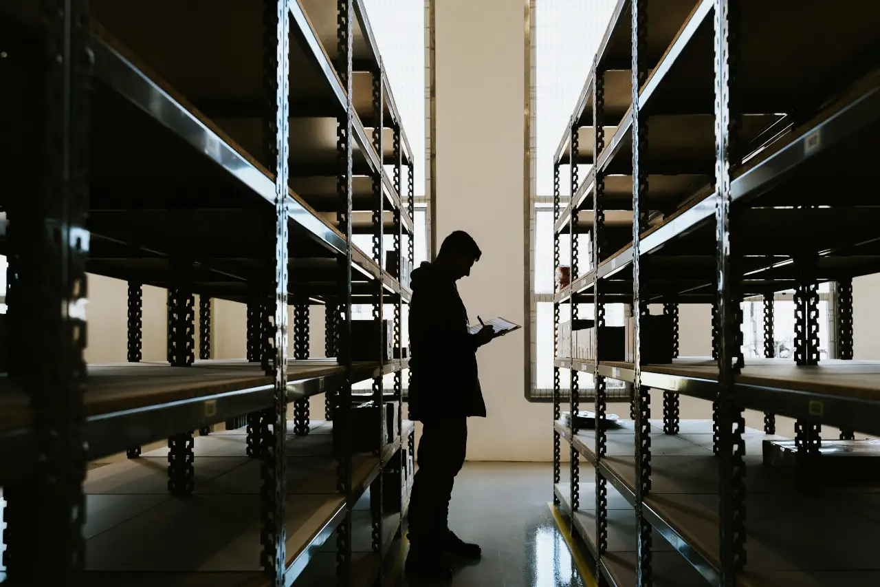 Silhouette of a person with a clipboard between warehouse shelves.
